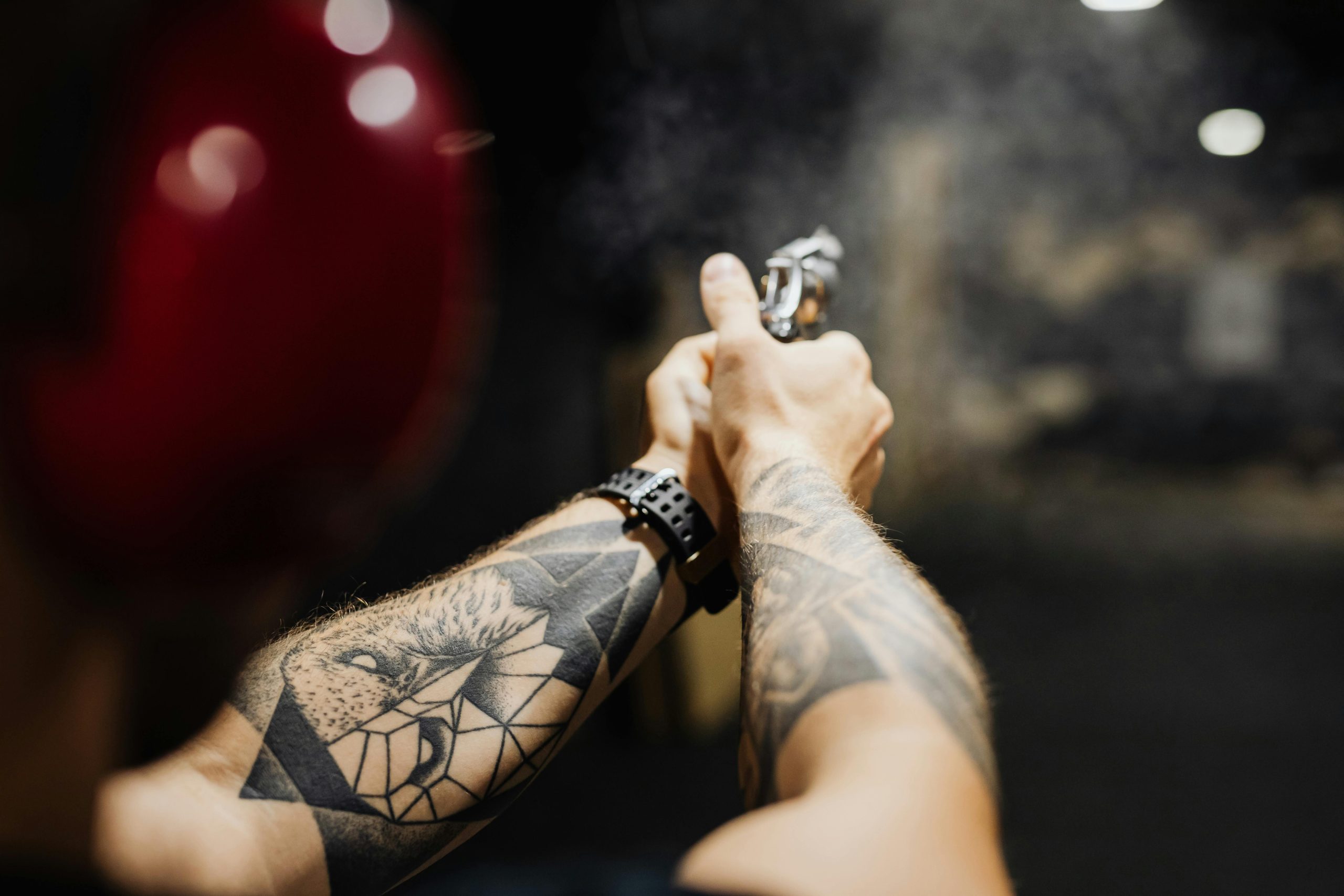 Close-up of tattooed arms aiming a gun at an indoor firing range with visible smoke.