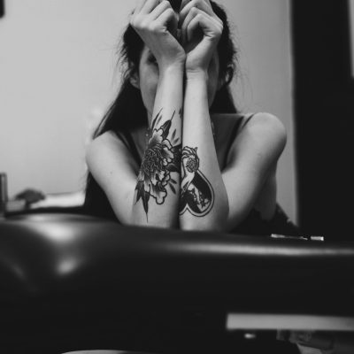 An artistic black and white portrait of a tattooed woman, arms raised, posing indoors.